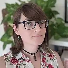 A white woman with brown hair, ear piercings, and thick plastic glasses wears a white, sleeveless shirt with a rose pattern; in the background is a green plant