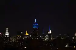 Upper floors of the Empire State Building lit in blue, amid other lit skyscrapers