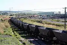 An empty freight train curving through an industrial complex, with a rural background of fields and hills