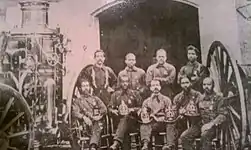 Nine men sitting between engine wheels as tall as they are