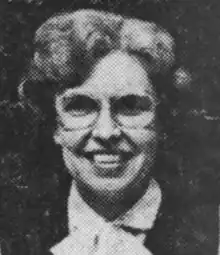 A smiling white woman with a set hairstyle and glasses, wearing a white blouse and a dark vest