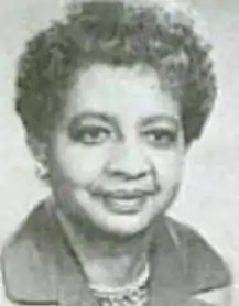 A smiling middle-aged Black woman with short curly hair, wearing a jacket with a wide dark collar