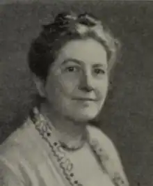 A white woman with hair in an updo, wearing glasses and a soft light-colored jacket