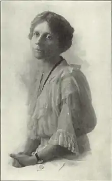 [In black and white] A woman seated with cropped or pulled back hair, wearing a white lacy dress, dark banded watch and dark ribbon or necklace. She faces the viewer.