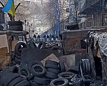 Photograph of security patrols standing behind a makeshift barricade of tires, burned vehicles, cardboard, and wooden pallets.