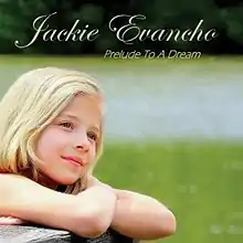 A photo of nine-year-old Jackie Evancho gazing over a grassy area and resting her crossed arms on a wooden fence railing.  A lake and wooded area are in the background.