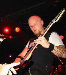 A bald man wearing a white shirt under a black vest playing a guitar