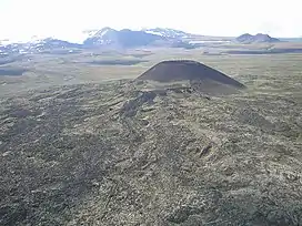 A barren plateau with a small cone-shaped mound in the foreground, a glaciated mountain in the left background and a small elongated mound in the right background.