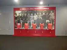 Five different Benfica shirts are displayed on a panel titled "HÁ MAIS DE 100 ANOS A VESTIR ESTA CAMISOLA" ("wearing this shirt for over 100 years") with a background picture of twelve Benfica footballers casually dressed and lined up.