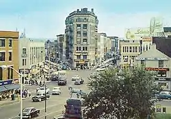 A view down on a busy city intersection with a tall wedge-shaped building at the junction of two streets that fork in the center. A tree and statue of a horse are at the bottom, in the foreground