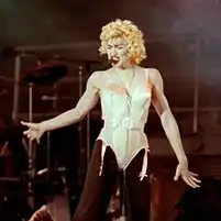 Madonna wearing a beige corset and black pants. She has blonde curly hair and has a headset microphone to her mouth.