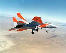 Jet aircraft with distinctive orange markings banking left over desert, with landing gears extended