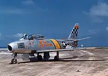 Glenn's silver fighter plane on the tarmac, with a yellow stripe behind the cockpit and a checkered pattern on the tail