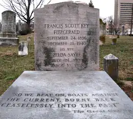 Photograph of the grave of F. Scott and Zelda Fitzgerald in Rockville, Maryland, taken during a snowless winter. The headstone reads: "Francis Scott Key Fitzgerald. September 24, 1896 - December 21, 1940. His wife Zelda Sayre. July 24, 1900 - March 10, 1948." Beneath the headstone is a gray slab inscribed with the final line of The Great Gatsby: "So we beat on, boats against the current, borne back ceaselessly into the past."