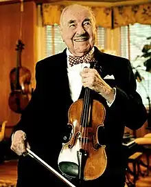 An older man wearing a suit and bow tie and smiling while holding a violin.
