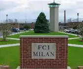 A red brick sign in the foreground says "FCI MILAN". In the back, a solitary green tree is visible atop a grassy mound, with a tower to its right.