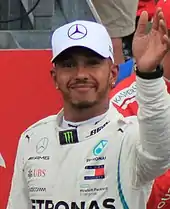 Lewis Hamilton wearing a silver baseball cap and racing outfit and waving to the crowd with his right hand