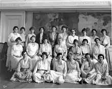 Photograph of 23 women arranged in three rows in front of a large painting depicting dancing women in diaphanous costumes. The first row of women are seated on the floor, the second row are seated in chairs, and the back row is standing.
