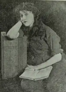 A young white woman with loose curly hair, seated, one arm resting on a crate, the other on a book in her lap.