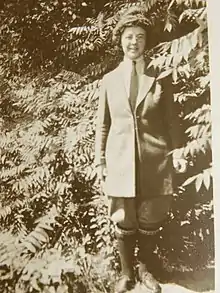 Picture of a woman standing outdoors, wearing jacket, pants, long socks and shoes, with trees and bushes close behind her