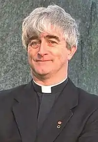 A middle-aged man in a priest's collar stands outside a house