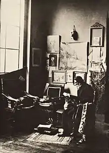 A seated man with a white beard reads a book at the corner of a studio. (cap)