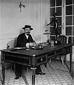 White man with neat imperial beard and moustache, sitting at a desk, in a straw hat