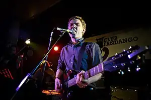 Peter Brewis of the rock band Field Music performing on a stage, playing a guitar and singing into a microphone, as other musicians perform in the darkened background behind him.