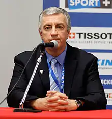 Man with grey hair sitting behind a microphone at a table, wearing a dark blue suit jacket, light blue dress shirt and striped grey necktie