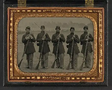 A sepia toned photograph of five soldiers standing at parade rest in a neat line