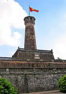 Stone tower on top of a stone wall. The wall has circular wheel-shaped windows and a red flag with yellow star is raised on top of the tower.