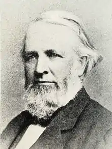 Head and shoulders photograph of a man in an overcoat.  He has a full beard with sideburns and has his white hair parted at the his right with it being of enough length to be tucked a few inches behind his left ear. He wears a serious expression and is looking to the left.