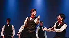 Four young men in tuxedoes sing on stage in front of a deep blue background. Two singers in particular stand in the foreground, gesticulating while singing a big note.