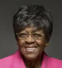 A headhsot of an older black woman who is smiling. She has short dark hair and is wearing a bright fuschia top