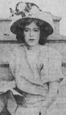 A young white woman with dark hair, wearing a light-colored dress and a brimmed hat