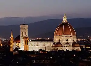 The Florence Cathedral, illuminated at night, showing the large red brick dome, a decorated white marble nave, and a vertical, white marble bell tower to the left. Mountains are visible in the background and a dark, low-lying city in the foreground.