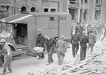 A casualty is carried by Civil Defence stretcher-bearers past an Austin K2/Y of the American Ambulance Great Britain following a V-1 flying bomb attack in London, 1944.