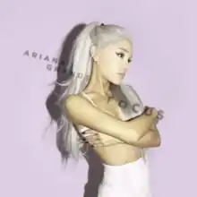 A picture showing a woman in a purple background with her arms through her body, her long ponytailed, platinum hair falls over her right shoulder. She wears a white mini skirt and a top.