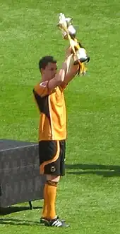 A young, dark-haired man in old-gold and black football attire stands with an ornate trophy raised in front of him.