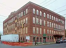 A four-story square brick building with boarded-up windows. There is a white trailer on its left and some signs from a construction company, both in a fenced-off area