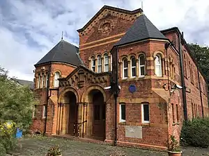 Dark red brick building with twin central entrance arches and central higher roof section flanked by projecting double towers against a dappled sky