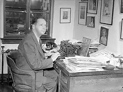 A black and white photograph of Foster Hewitt sitting on a chair in front of a desk