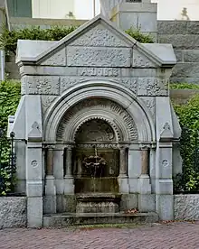 A small water fountain with a gothic structure along a brick sidewalk, flanked by wrought iron fences