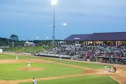 A view of the green baseball field from the third base side seats showing men in white baseball uniforms playing their positions at dusk
