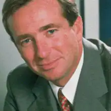 The image shows a brown-haired man in a suit. He has a slight smile and is looking into the camera.
