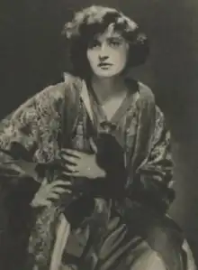 A young white woman with bobbed dark hair, wearing an oversized print robe, one hand on hip, one on chest