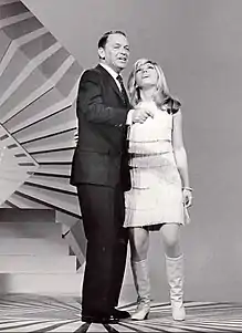 A dark-haired man in a suit and a younger blonde woman in a short dress singing together