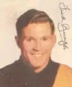 A red headed man smiles in portrait while wearing a black football jumper with a yellow sash