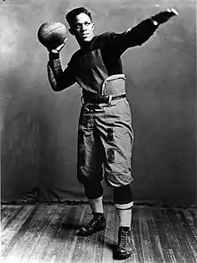 Pollard in football uniform staring off in the distance and striking a classic football pose; one arm is outstretched, while the other holds a football, as if he were about to throw it.