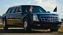 A black Cadillac limousine is at a 45-degree angle, showing to the camera its front and starboard sides.  Two small flagpoles are mounted to the front fenders, one flying a US flag with gold fringe.